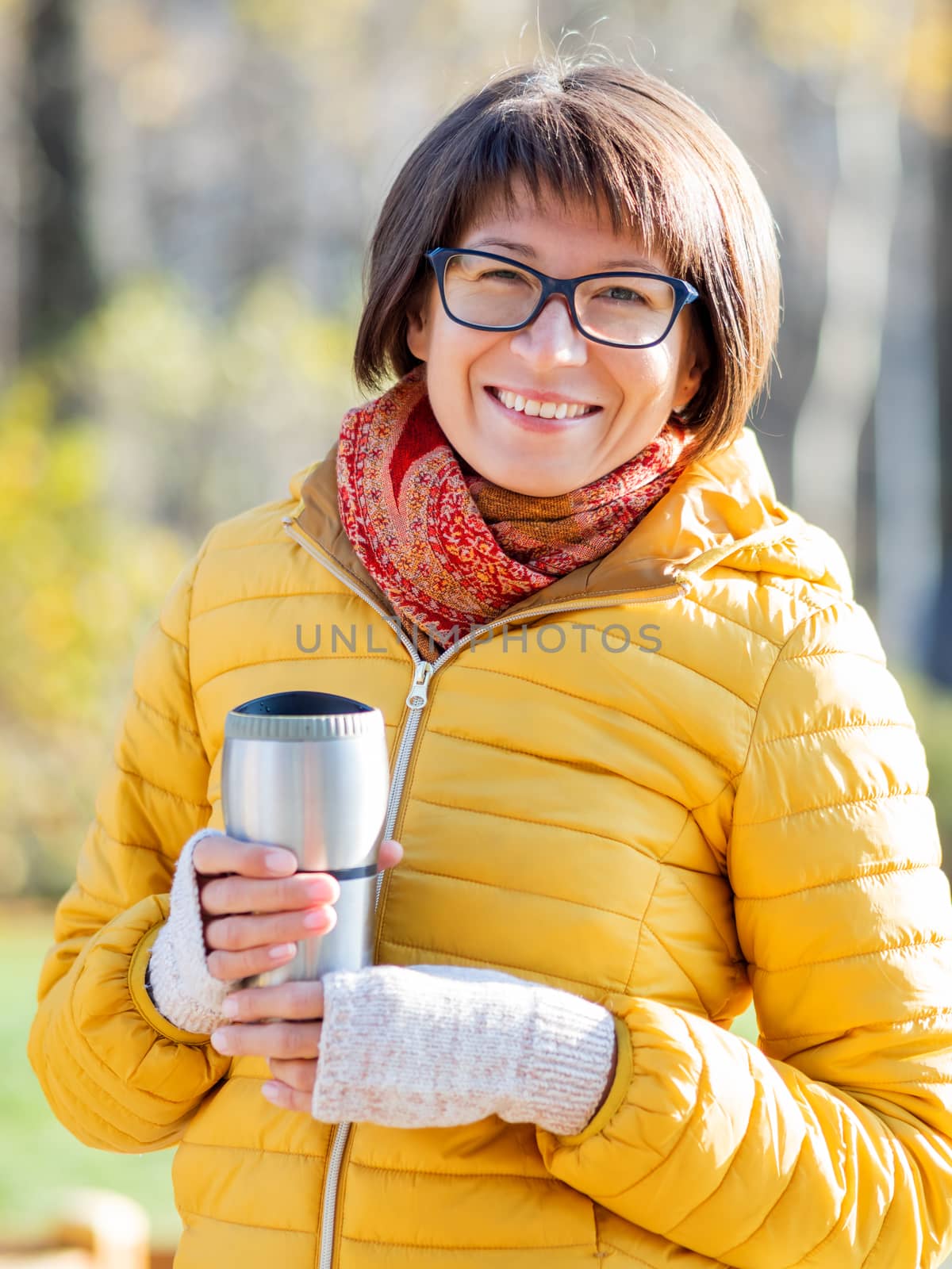 Happy wide smiling women in bright yellow jacketis holding thermos mug. Hot tea or other beverage on cool autumn day.