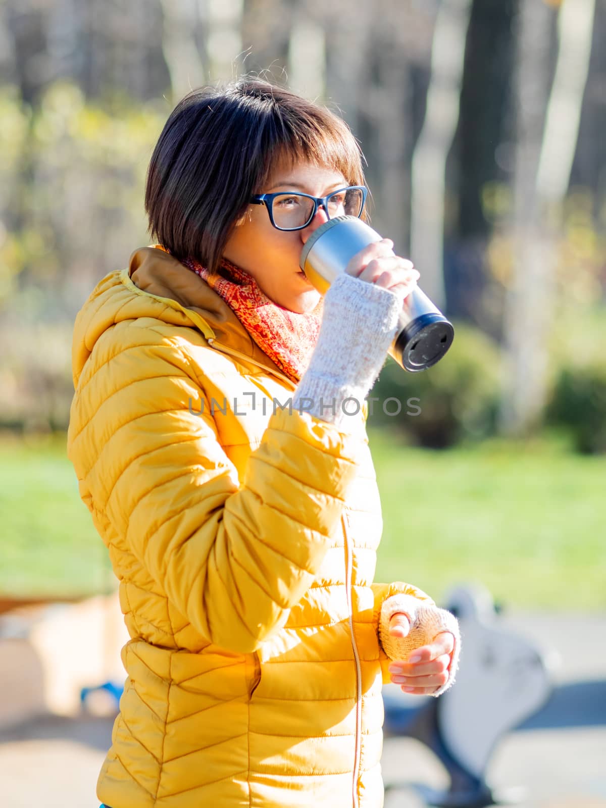 Happy wide smiling women in bright yellow jacket is holding thermos mug. Hot tea or other beverage on cool autumn day.