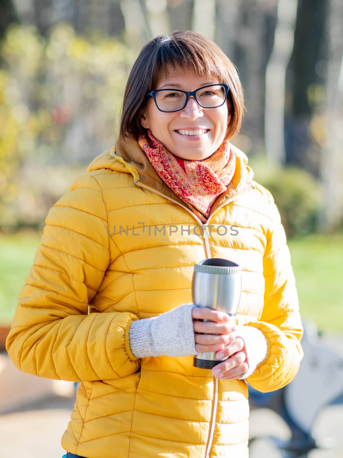 Happy wide smiling women in bright yellow jacket is holding thermos mug. Hot tea or other beverage on cool autumn day.