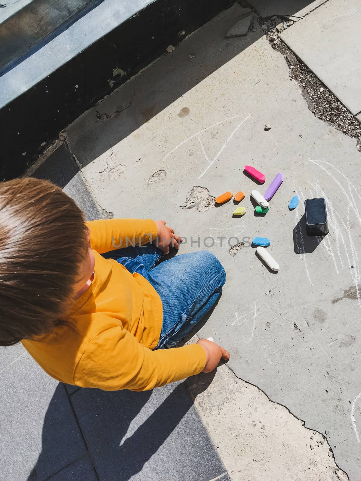 Toddler in jeans draws with crayons on the asphalt in sunny day. Child is holding colored crayons. Kid's hands and clothes are covered with colorful stains. Outdoor leisure activity.