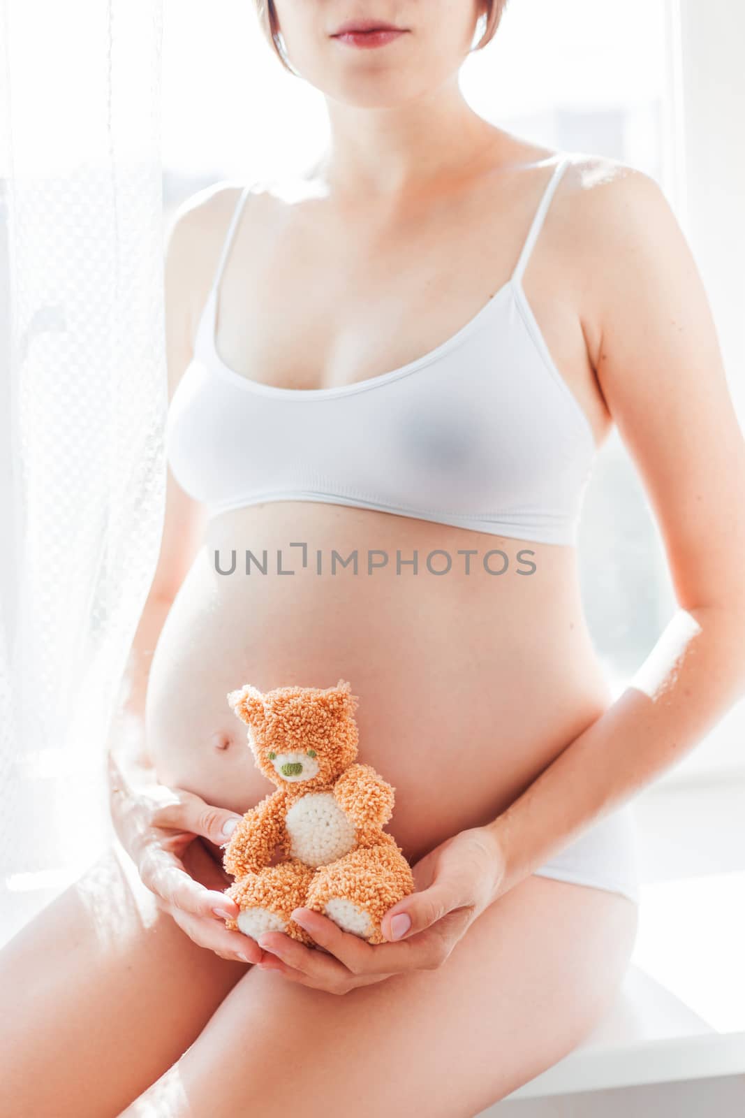Pregnant woman in white underwear with knitted toy teddy bear. Young woman expecting a baby. Cozy happy background in sunny morning.