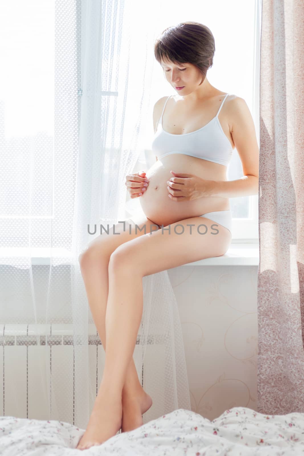 Pregnant woman in white underwear sitting on window sill. Young woman expecting a baby. Cozy happy background in sunny morning.