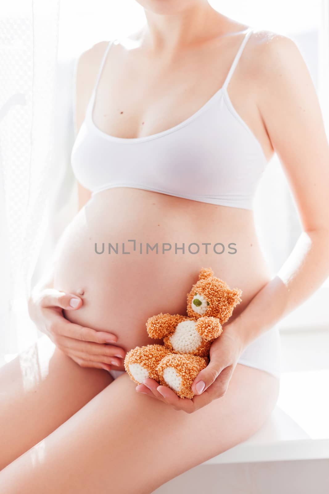 Pregnant woman in white underwear with knitted toy teddy bear. Young woman expecting a baby. Cozy happy background in sunny morning.