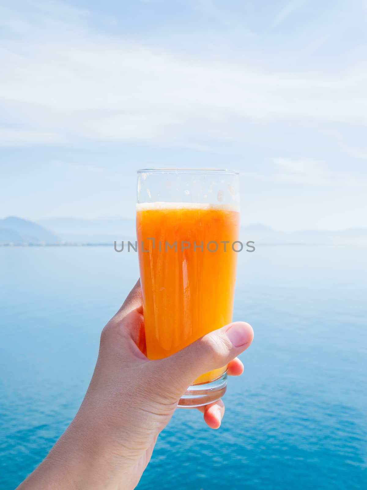 Hand with fresh tasty freshly-squeezed juice of ripe oranges in glass. Calm sea with mountains on the horizon. Kemer, Turkey. by aksenovko