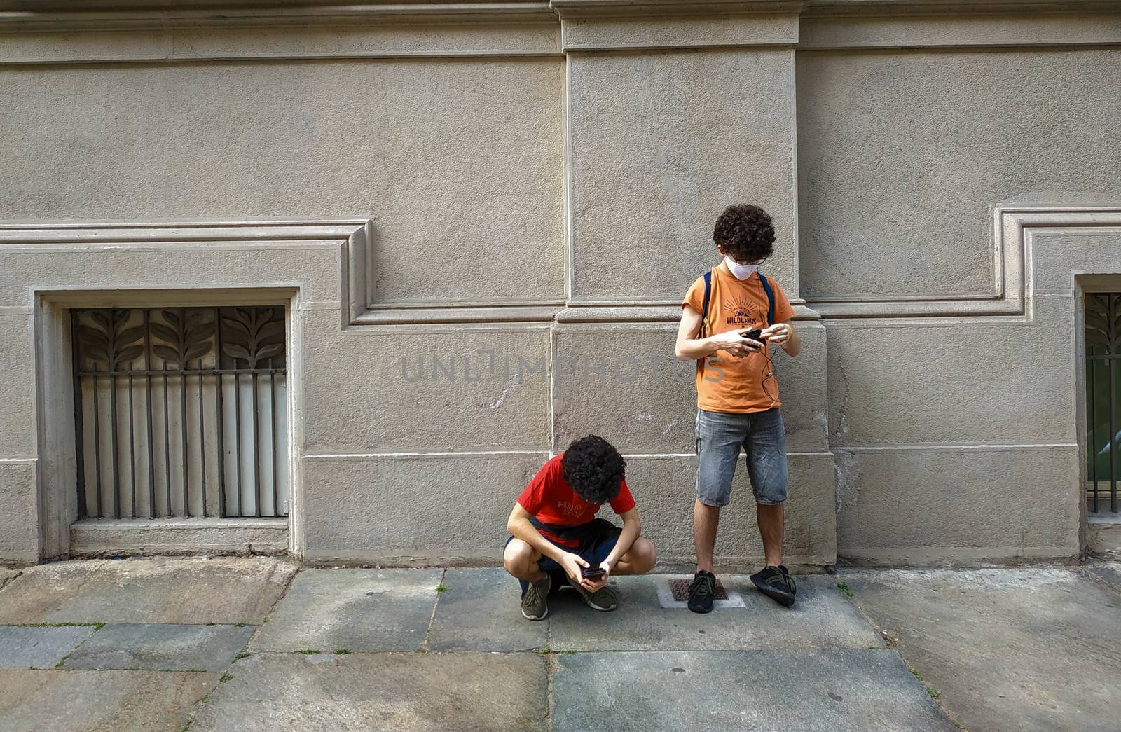 Two Caucasian boys use smartphones in the time of the coronavirus: one crouched and the other leaning against the wall. They wear a white protective mask.