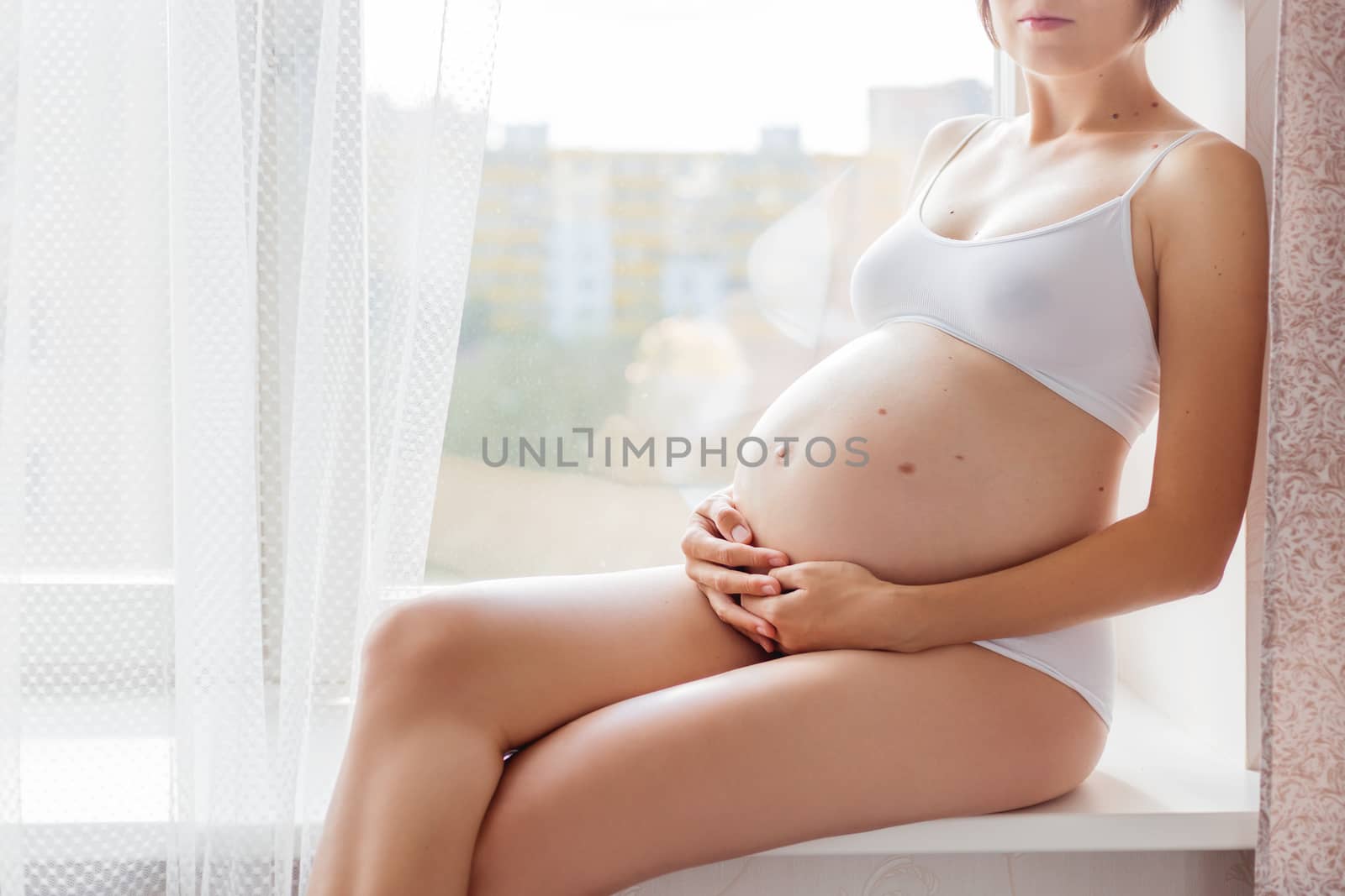 Pregnant woman in white underwear sitting on window sill. Young woman expecting a baby. Cozy happy background in sunny morning.