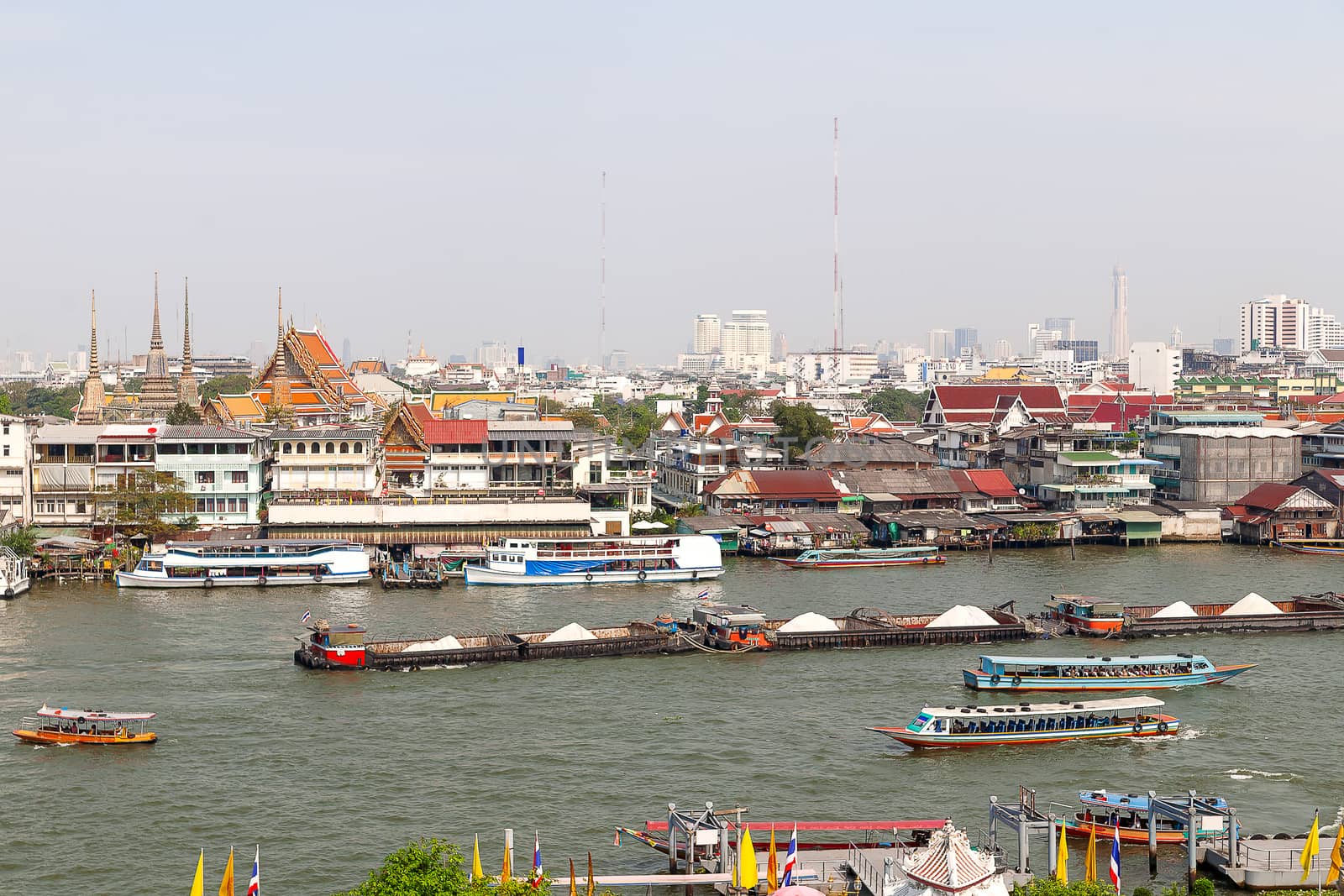 Colorful trasitional river boats crossing the Chao Phraya river in Bangkok, Thailand. With thai houses and skyscrapersi n the background. Cityscape in early misty morning.