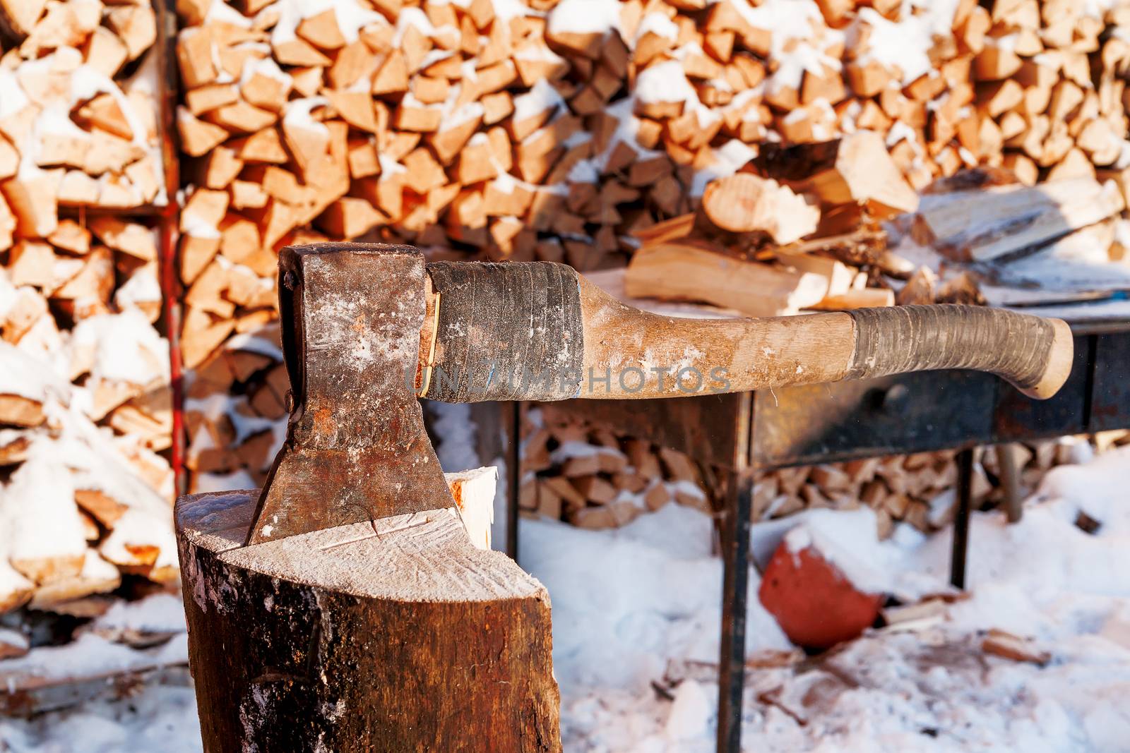 Axe in a wooden deck. Winter sunny day. Rural background with folded pile of firewood.