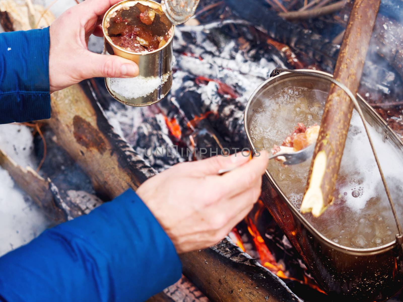 Cooking soup on a fire pot. Tourist puts stewed meat in soup from tin can. Winter camping in forest.