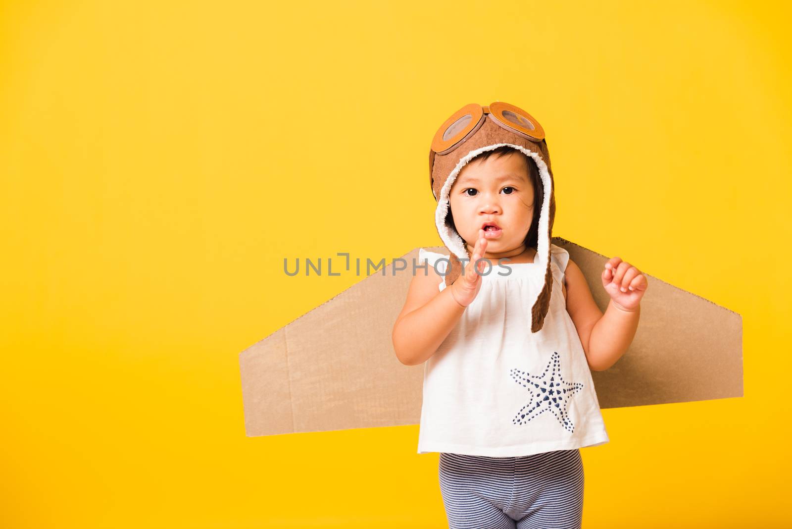Happy Asian beautiful funny baby little girl smile wear pilot hat playing and goggles with toy cardboard airplane wings flying, studio shot isolated yellow background, Startup freedom concept
