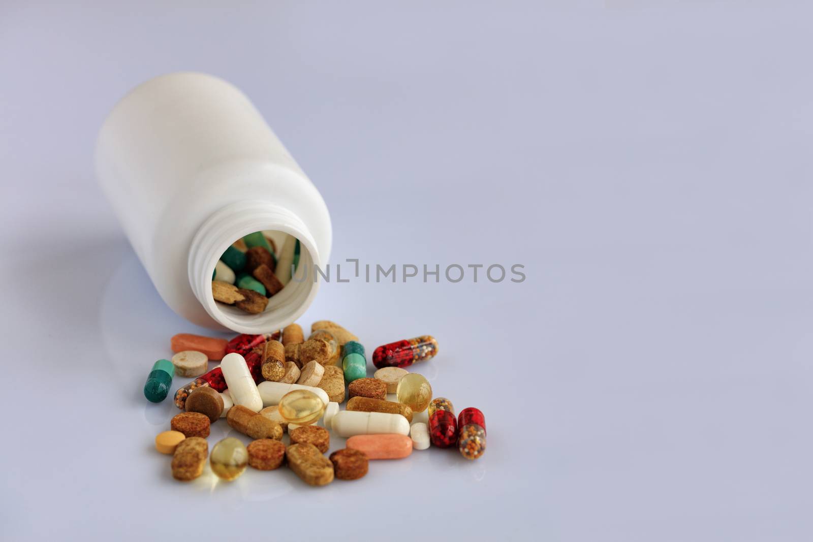 Tablets and capsules are scatter from a white bottle on a white smooth table.