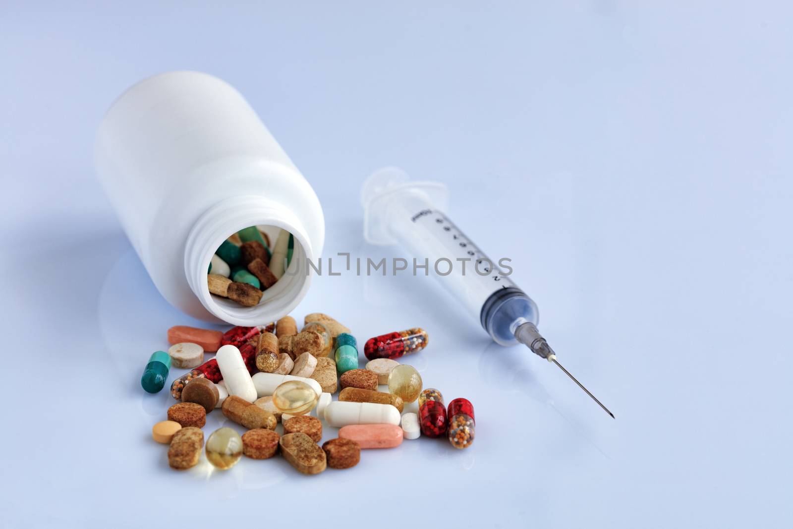 Tablets and capsules are scattered from a white bottle on a white smooth table near a disposable syringe.