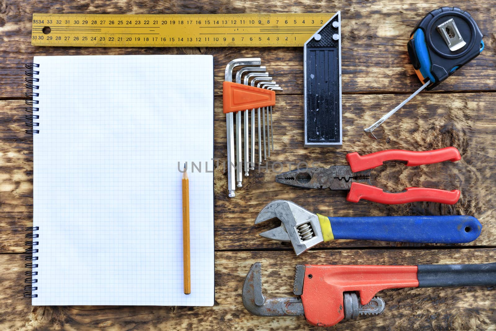 Old hand tools, pliers, tape measure, adjustable wrenches, a set of hex keys indicate an empty notebook with a pencil against the background of an old wooden table.