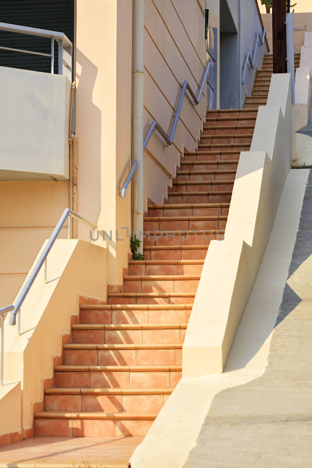 The path goes up a stone staircase from beige paving slabs in the bright sun in a narrow Greek street.