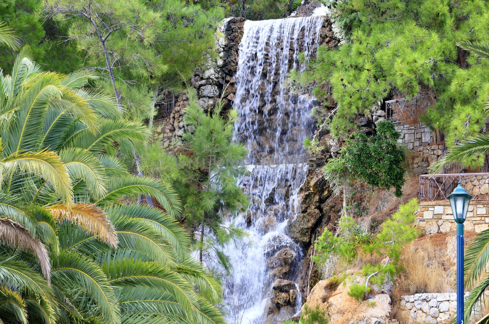 A large waterfall from a spring of radon water flows down large boulders at the foot of the mountain among the green Mediterranean pines and palm trees in the park of Loutraki, Greece.