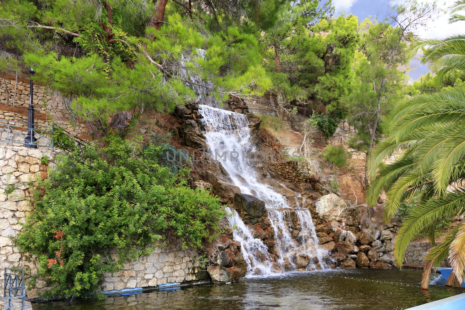 A large waterfall from a spring of radon water flows down large boulders at the foot of the mountain among the green Mediterranean pines and palm trees in the park of Loutraki, Greece.