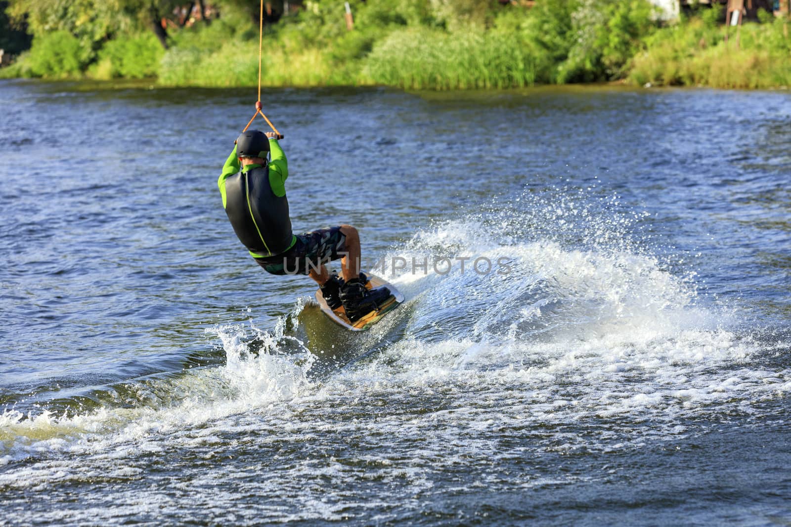 A wakeboarder rushes through the water along the riverbank, holding the cable with both hands and leaving a waterway behind him.