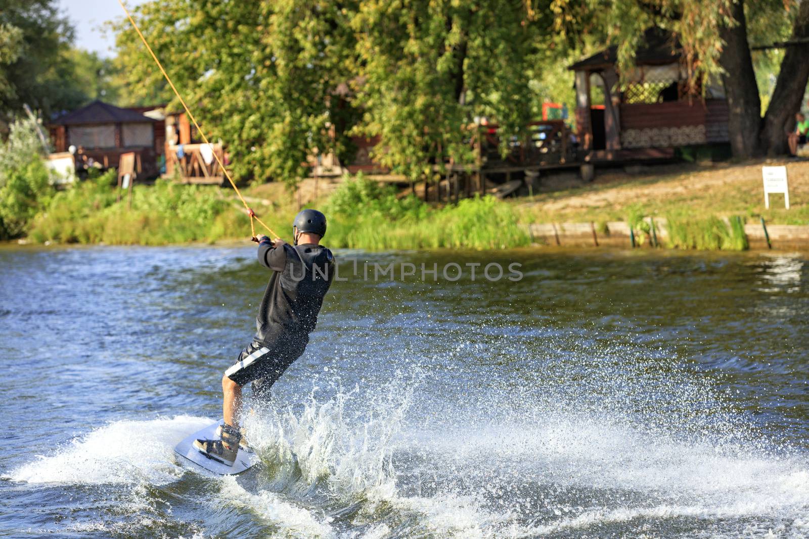 A wakeboarder rushes through the water along the riverbank, holding the cable with both hands and leaving behind a spray of water and a seething trail.