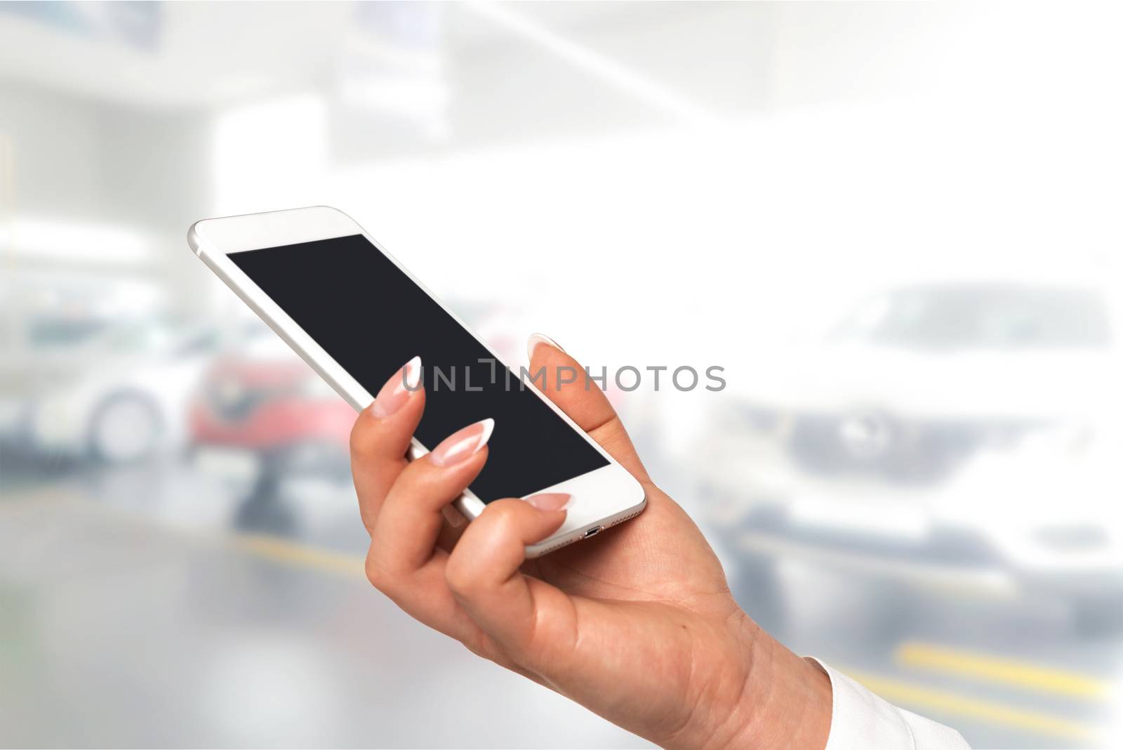 Smart car concept - female hand with perfect fingernails holding smartphone with blank screen on a blurred background of the auto dealership.