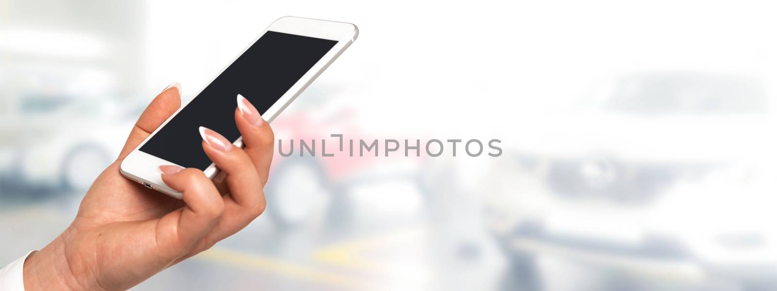 Smart car concept banner - female hand  holding smartphone with blank screen on a blurred background of the auto dealership (copy space).
