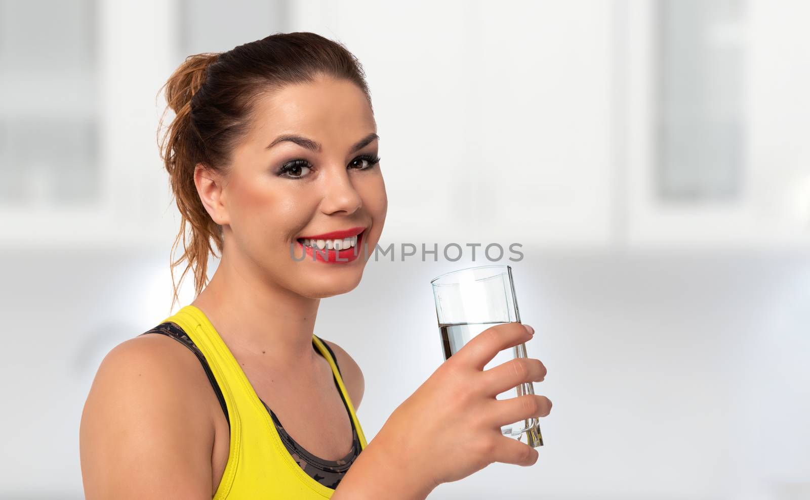 Healthy life concept - beautiful and young woman holds glass with fresh water on a blurred background of the kitchen (copy space).