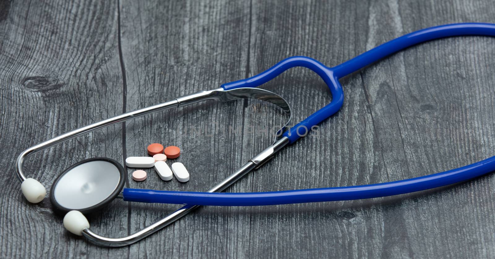 A blue medical stethoscope and a variety of medical pills sit on a wooden table.