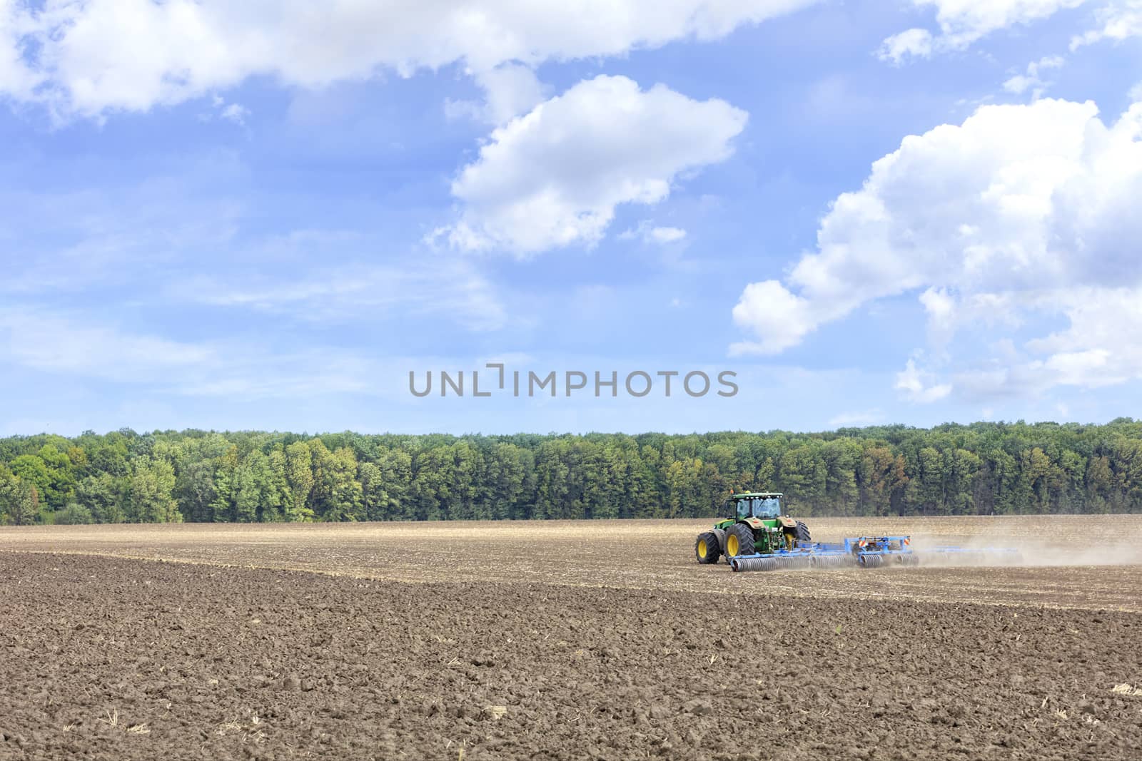 A tractor in a field shallowly plows the soil with metal discs after harvesting against a blue cloudy sky.