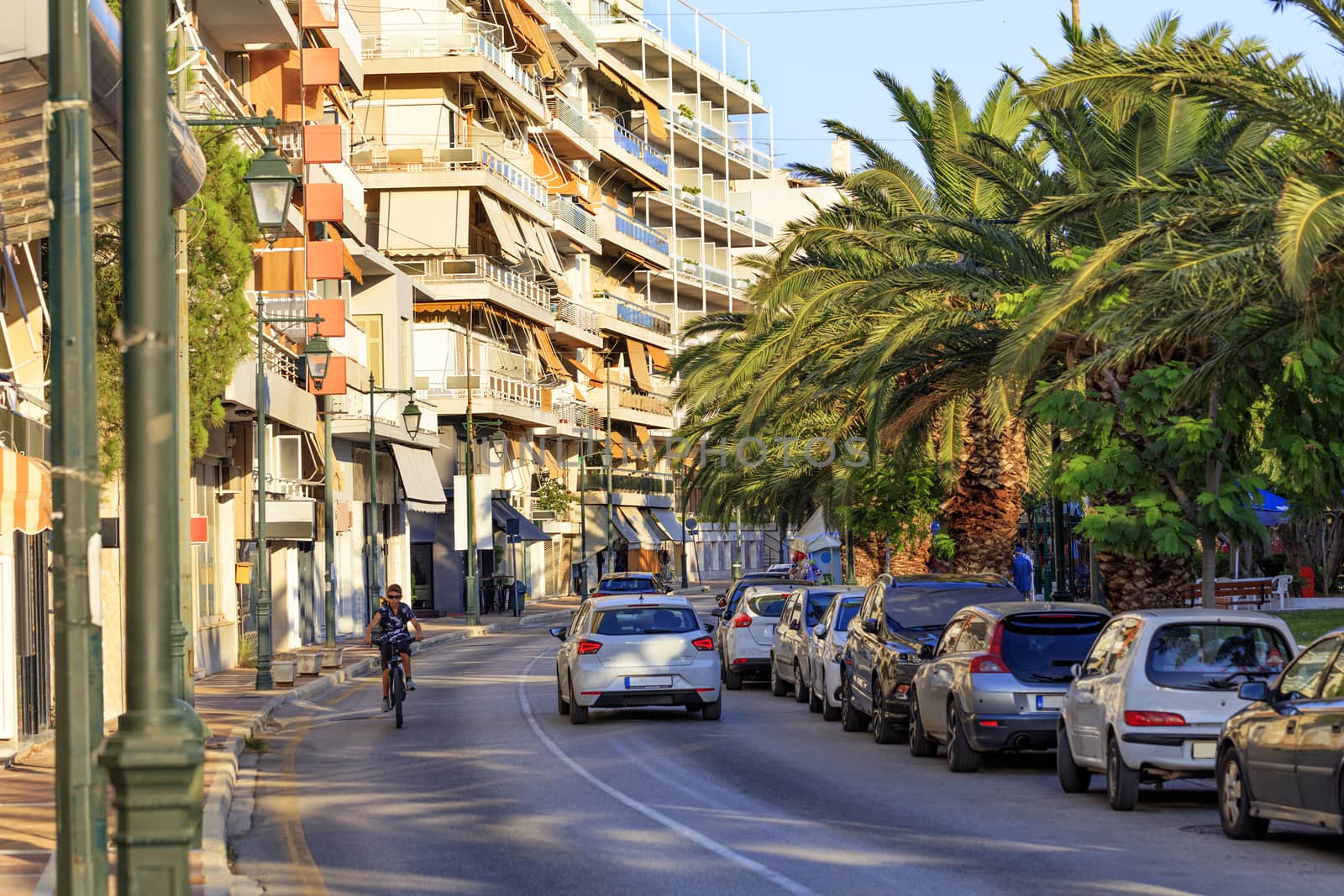 The asphalt road of the street of Loutraki city, which is flooded with sunlight and a teenager rides a bicycle on the road, the sky is reflected in blue light on the roofs of passing cars in the shade of spreading palm trees.