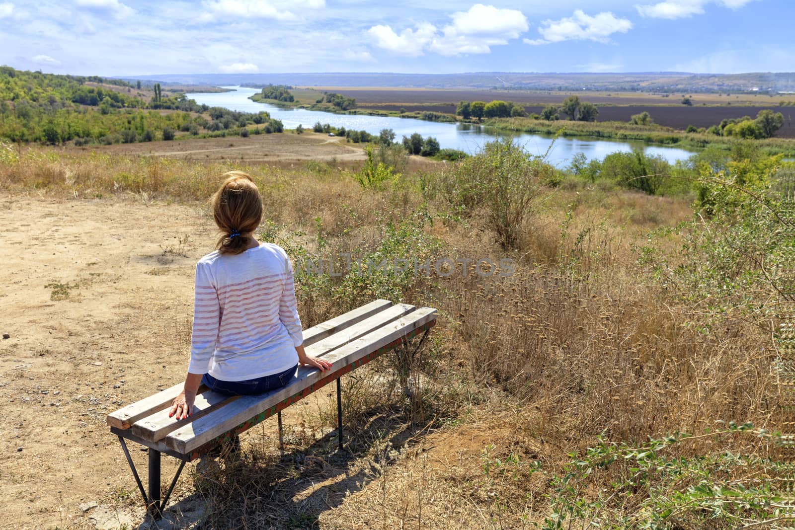 A young woman in the rays of the autumn rising sun greets the dawn on the banks of the Southern Bug River, sits on a wooden bench and looks into the distance.