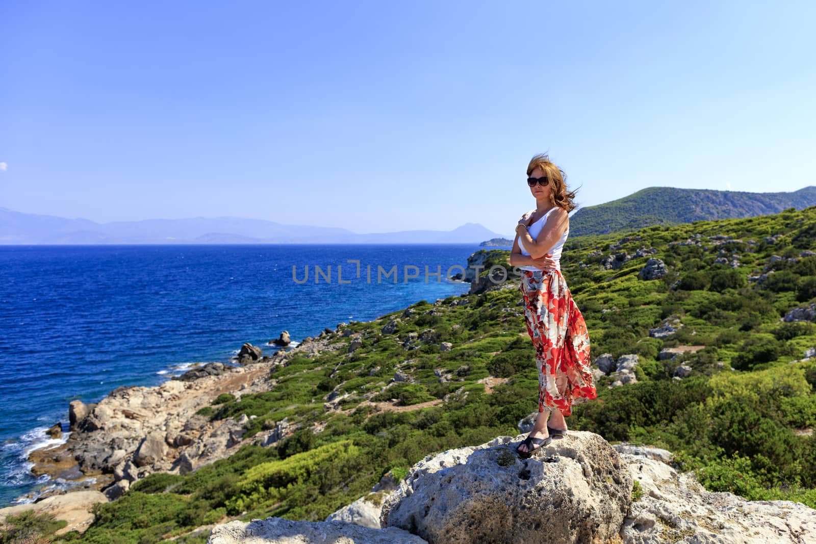 A beautiful lady stands on a large stone boulder against the background of the coastline of the Ionian Sea. by Sergii