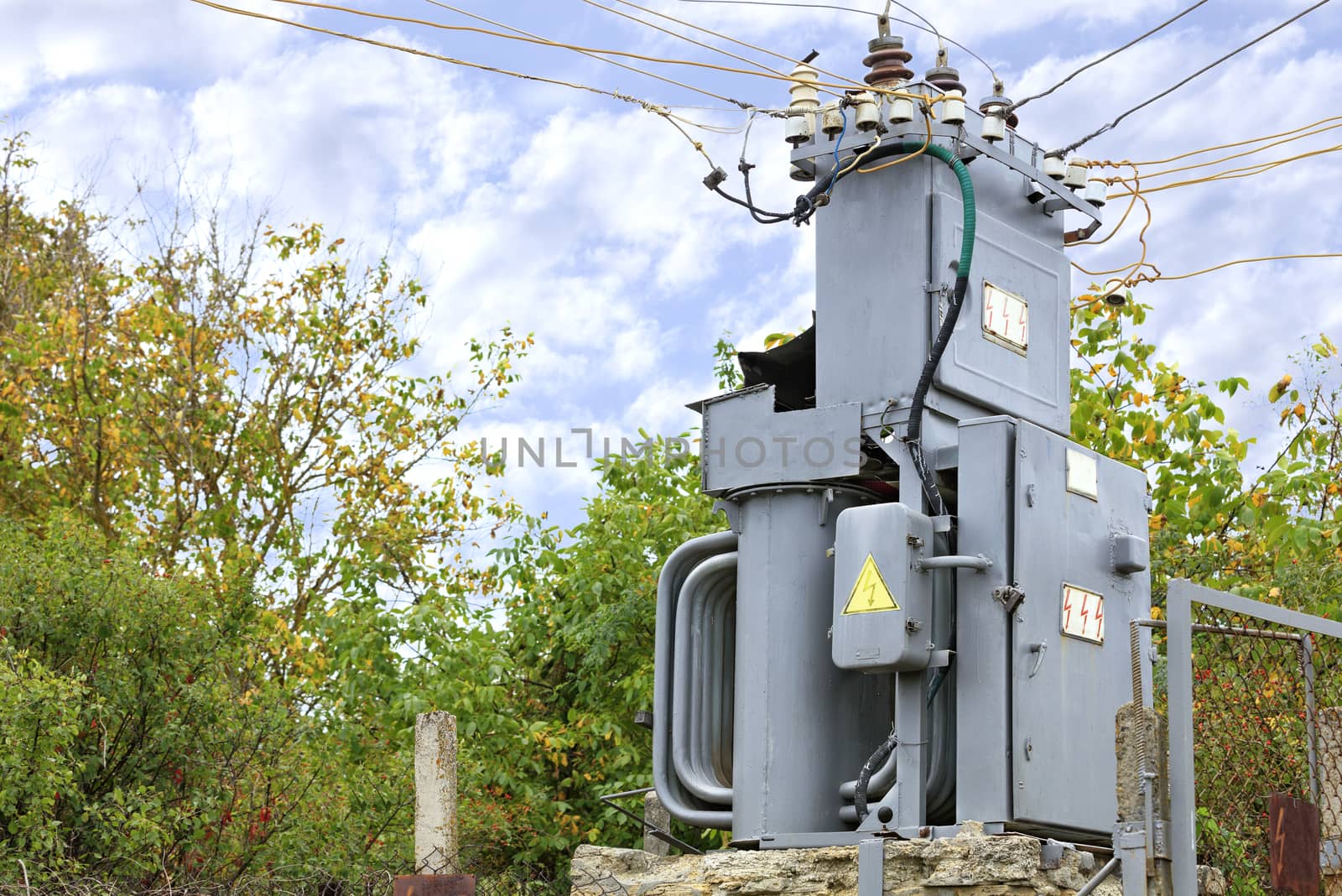 An old transformer substation stands in the middle of an autumn garden fenced with a metal mesh against a blue cloudy sky