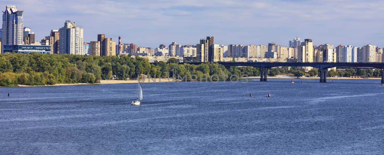 Sailing yacht goes along the banks of the Dnipro River against the backdrop of urban residential areas and a bridge over the river.