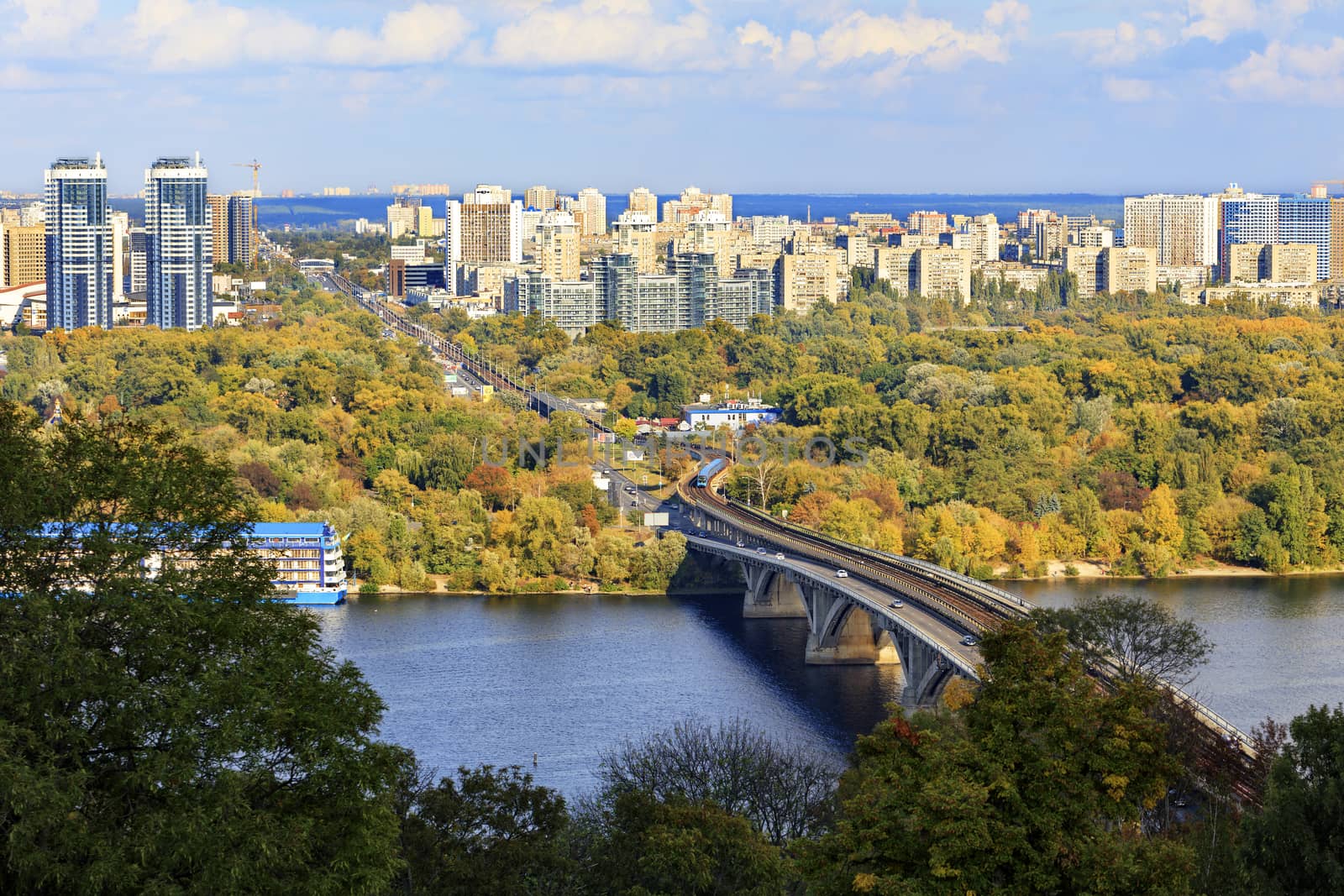 Autumn colors of the cityscape of Kyiv overlooking the Dnipro River, the railway line and the metro bridge, Hydropark and the left bank of the Rusanovka district under bright sunlight.