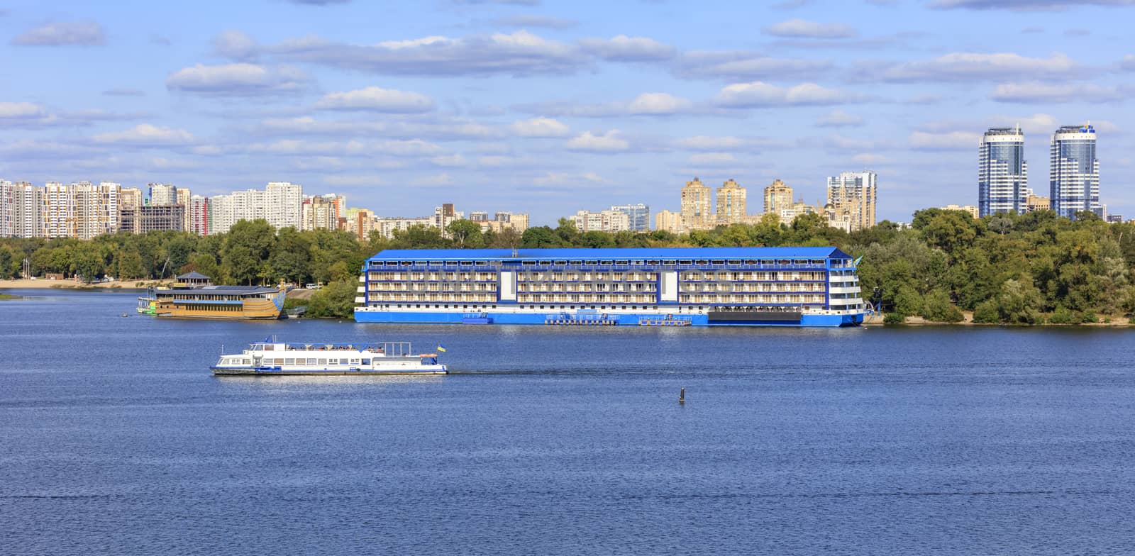 Pleasure boats and yachts go along the Dnipro River along the banks against the backdrop of urban residential areas of Kyiv on a bright sunny day.