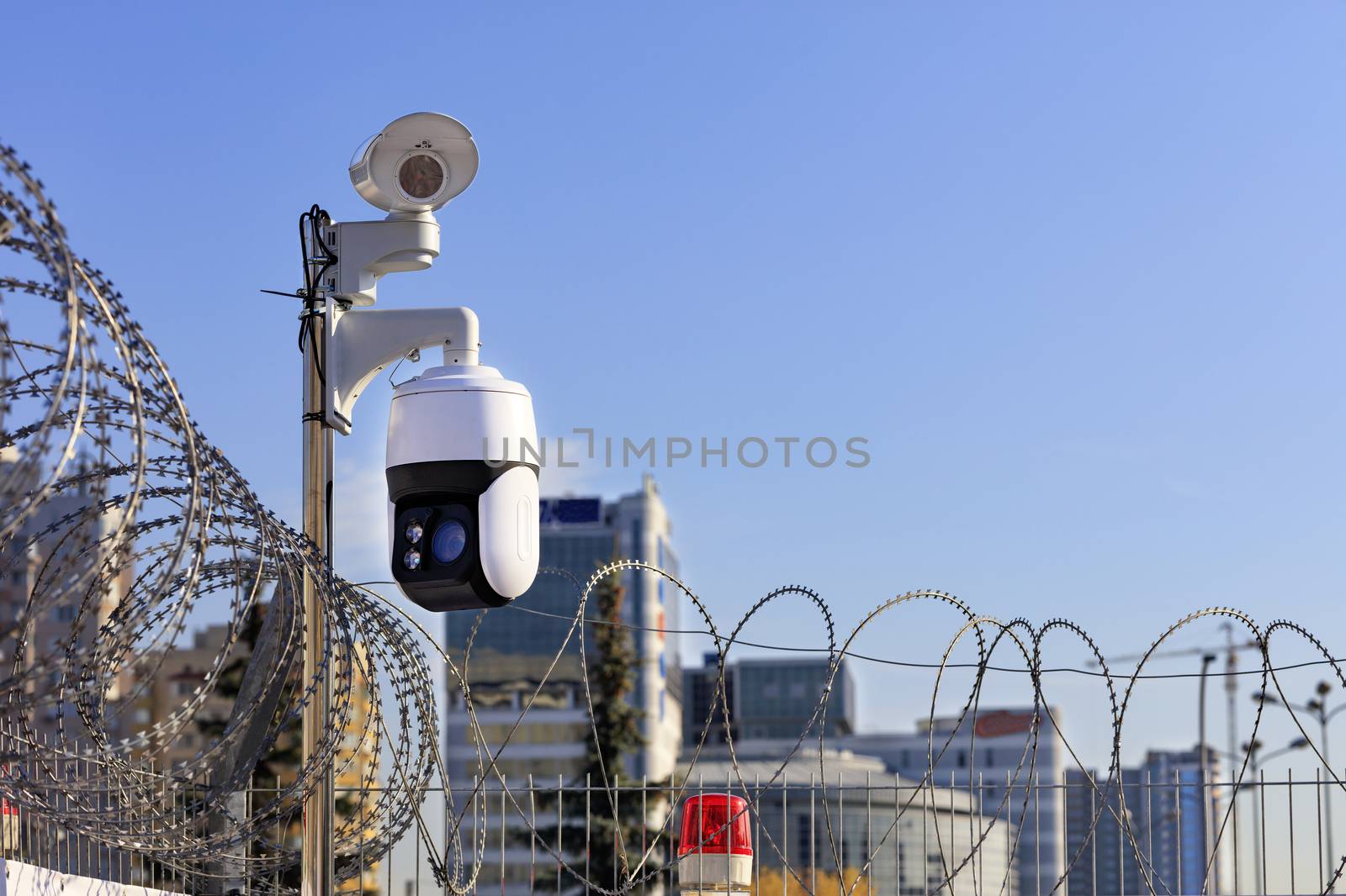 Security perimeter. Barbed wire fencing, a red light and cameras protect against intrusions against the background of urban buildings and the blue sky in blur.