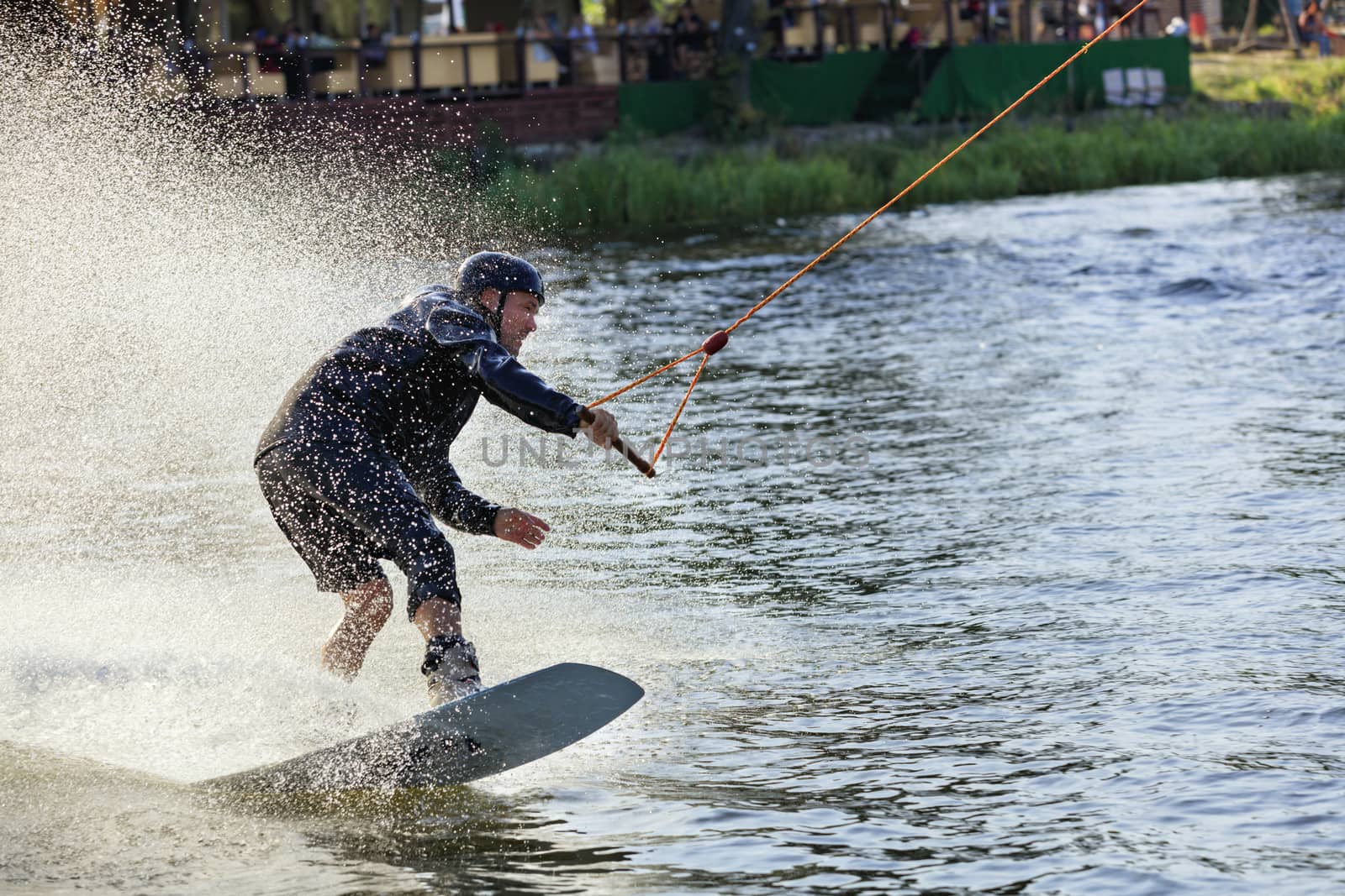 Wakeboarder rushes through the water along the grassy banks of the river, holding the cable with one hand and creating a cloud of spray behind himself.