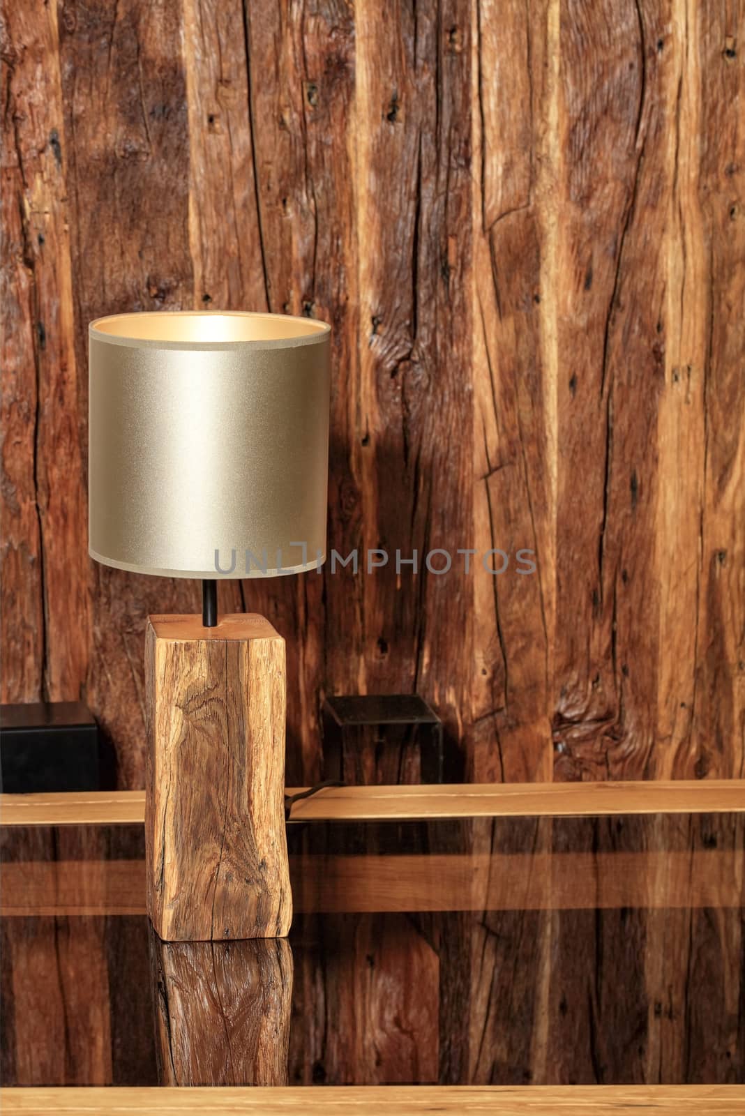 Beautiful modern lamp made of old wood in the form of a table lamp stands and is reflected on a polished wooden table against the background of an old cracked wooden interior.