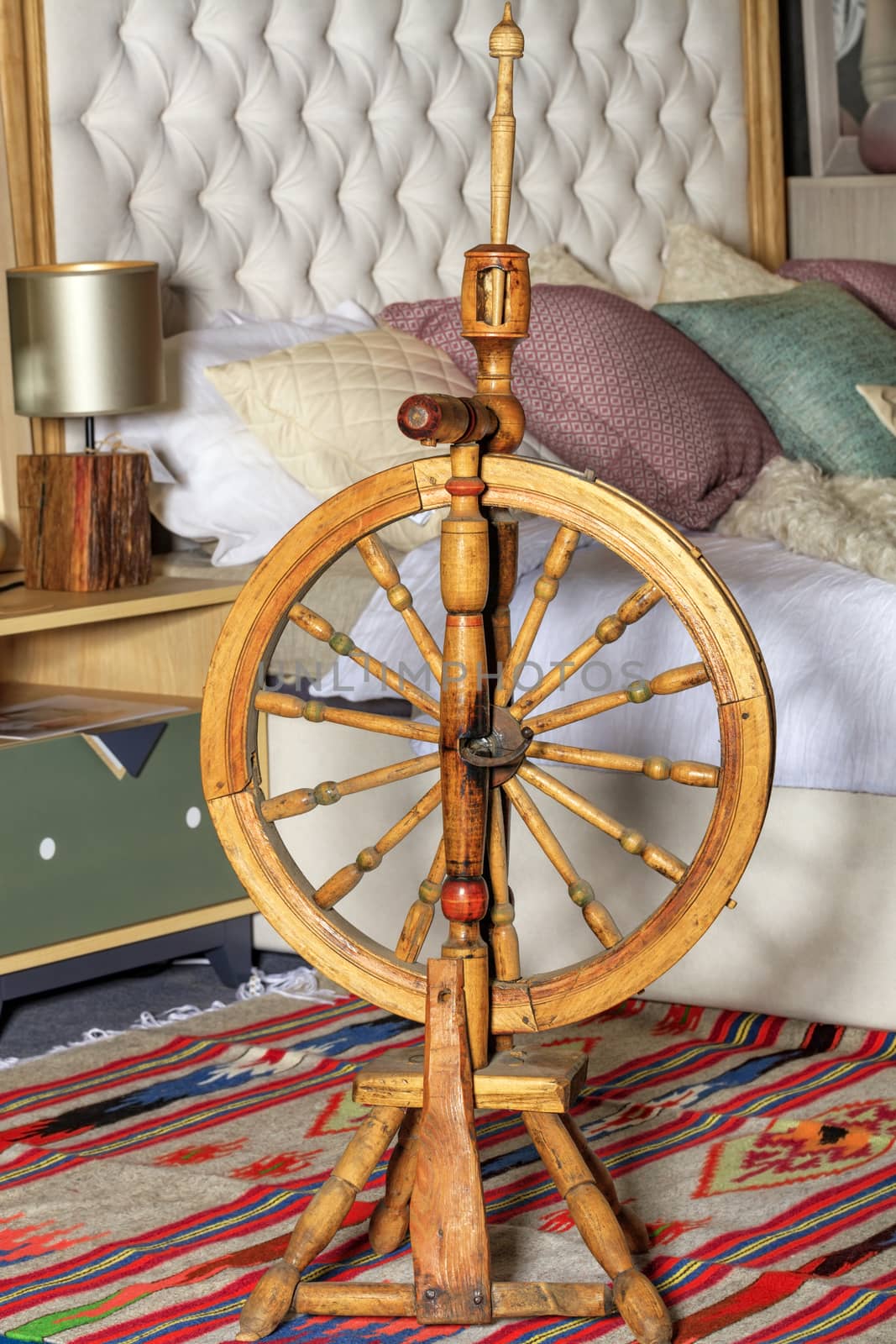 An old-fashioned spinning wheel stands on a homespun carpet at the head of the bed in the bedroom as part of an old rustic decor.