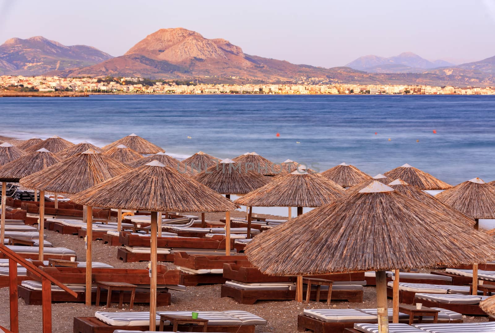 The straw tops of beach umbrellas and wooden deck chairs with mattresses on the deserted beach promenade in the rays of the evening setting sun. by Sergii