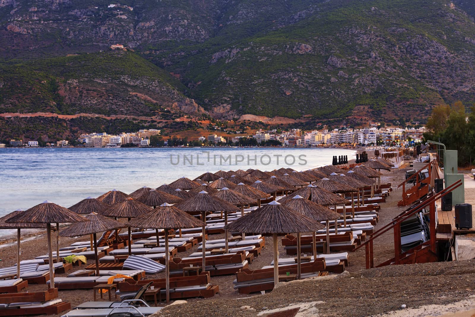 Thatched peaks of beach umbrellas with deck chairs on the deserted shore of the sea promenade against the background of evening red twilight light and the rays of the setting sun, mountain ranges in haze and blur with the city of Loutraki in Greece on the horizon.