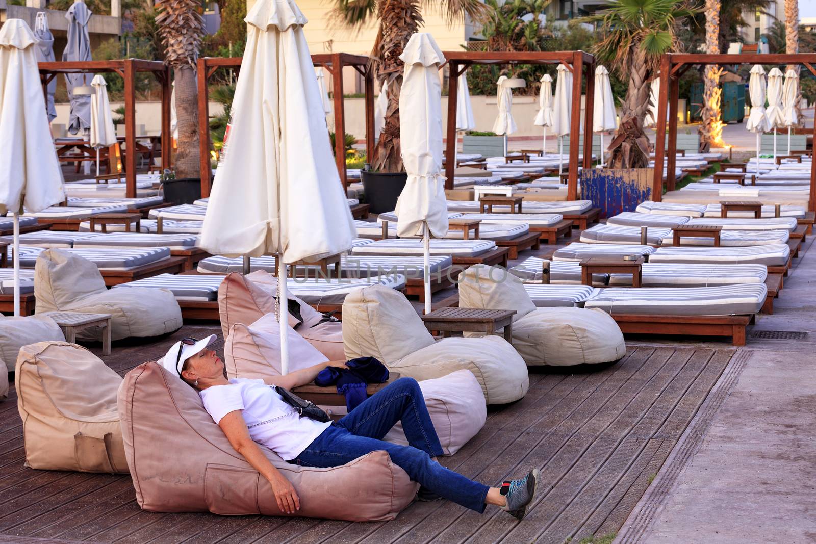 Tired middle-aged traveler is resting on a beach pillow among the collected beach umbrellas with sun loungers under palm trees on an empty beach amid evening red twilight light and the setting sun with copy space.