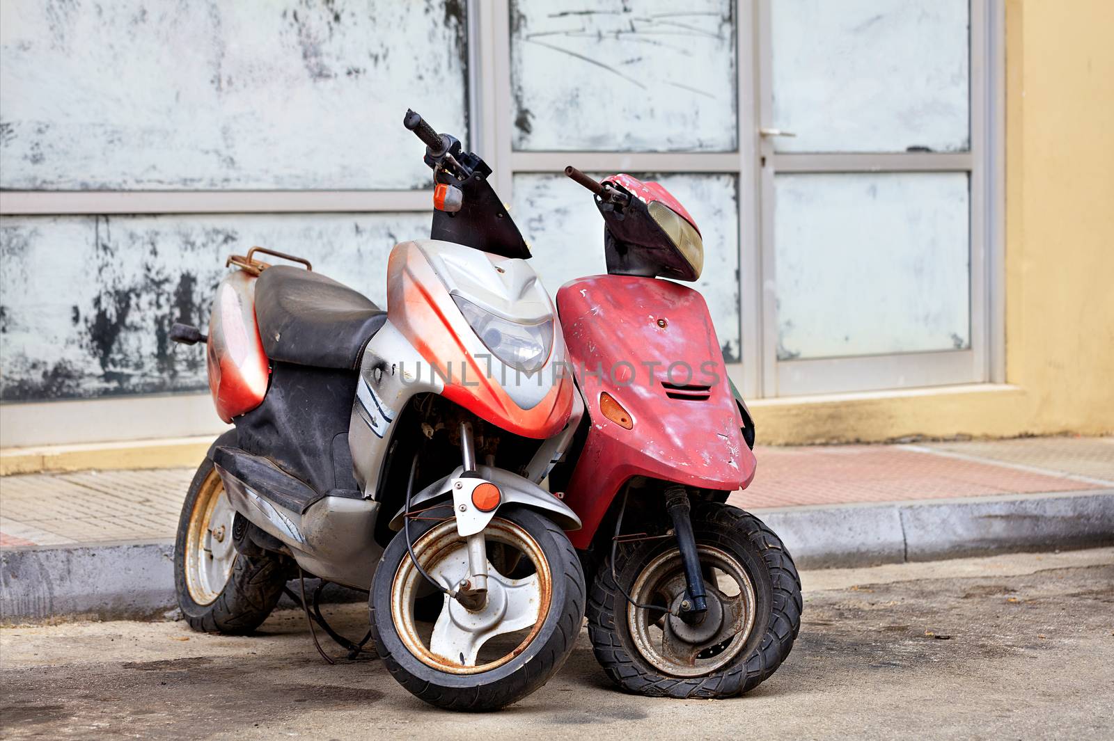 Two old worn scooters, red and silver, stand alone on the side of the road against the background of an old shop window under construction painted in white paint.