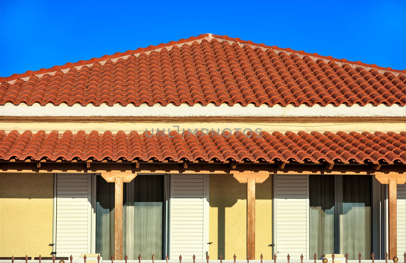 Sloped light brown clay tile roof of a rural traditional one-story house in southern Greece is covered with brown tiles against a blue sky.