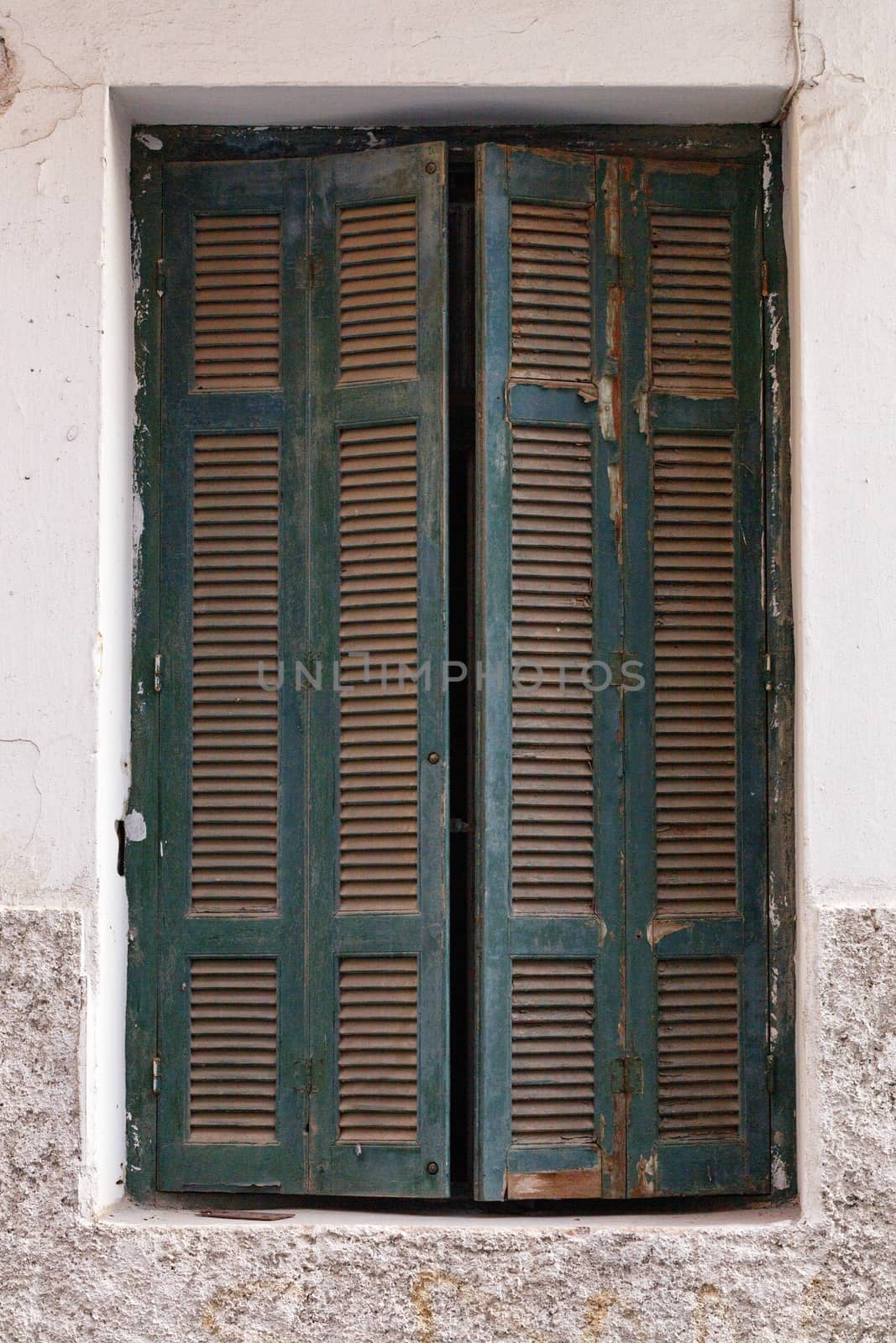 Old green wooden shutters are ajar on the old window against the background of the facade of the house.