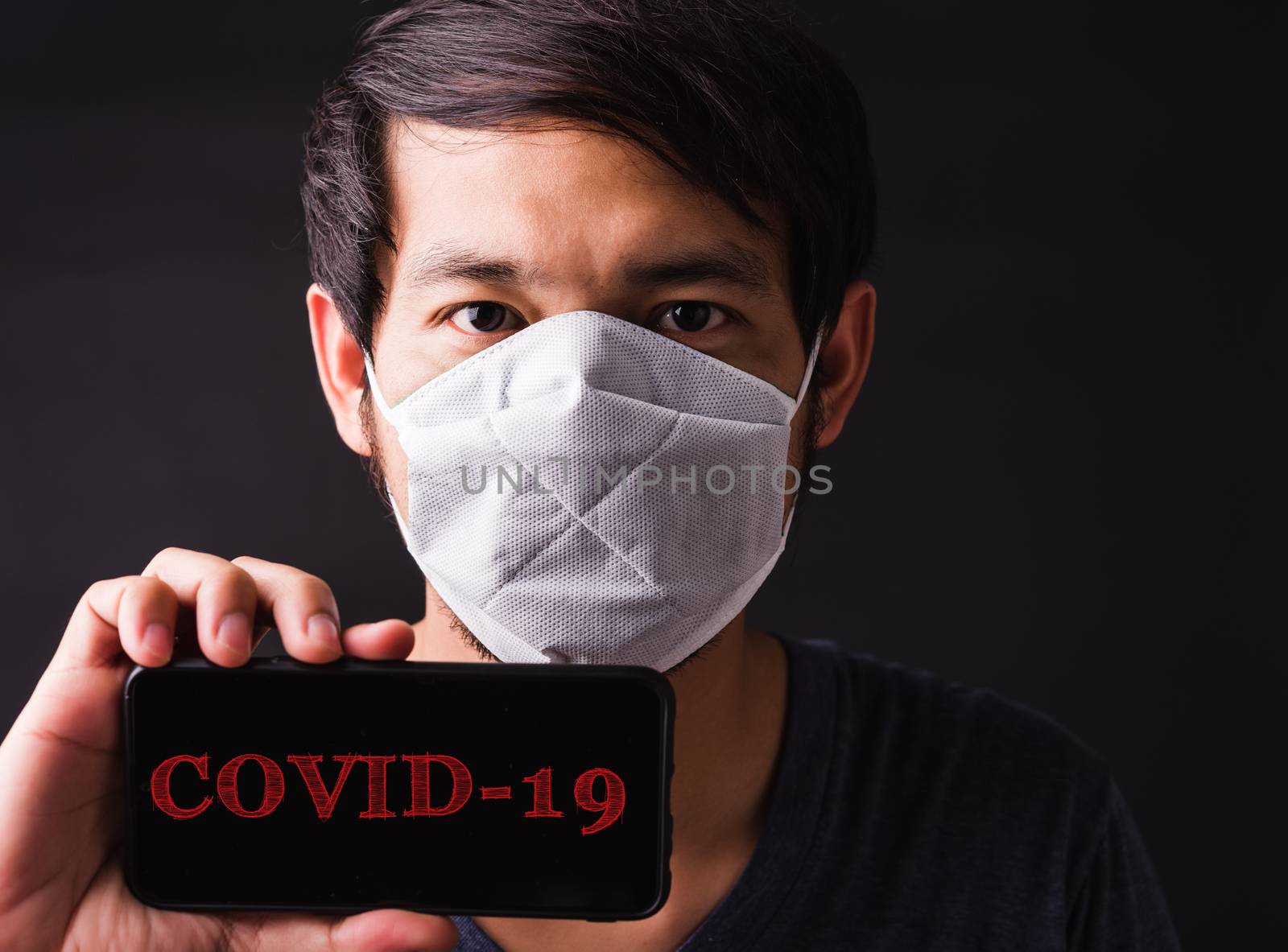 Closeup Asian handsome man wearing protective face mask fear in eye holding show mobile phone blank screen near face, hygiene prevention COVID-19 or coronavirus protection concept, black background