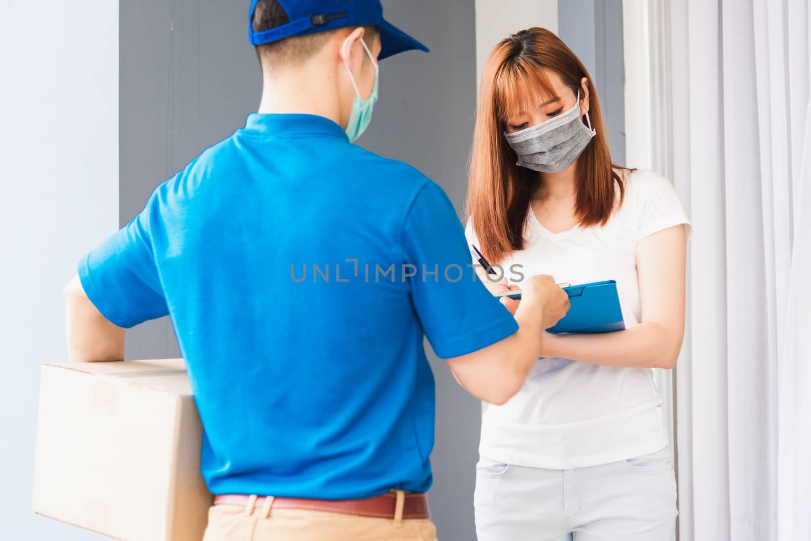 Asian young delivery man courier shopping online with package post box in uniform protective face mask coronavirus he making service woman customer using pen signature on paper book at home, COVID-19