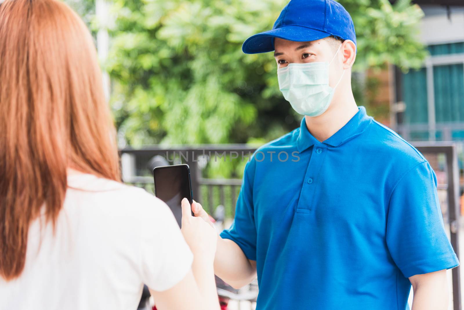 Asian delivery express courier young man giving parcel boxes to woman customer signature for receiving on mobile phone both protective face mask, under curfew quarantine pandemic coronavirus COVID-19