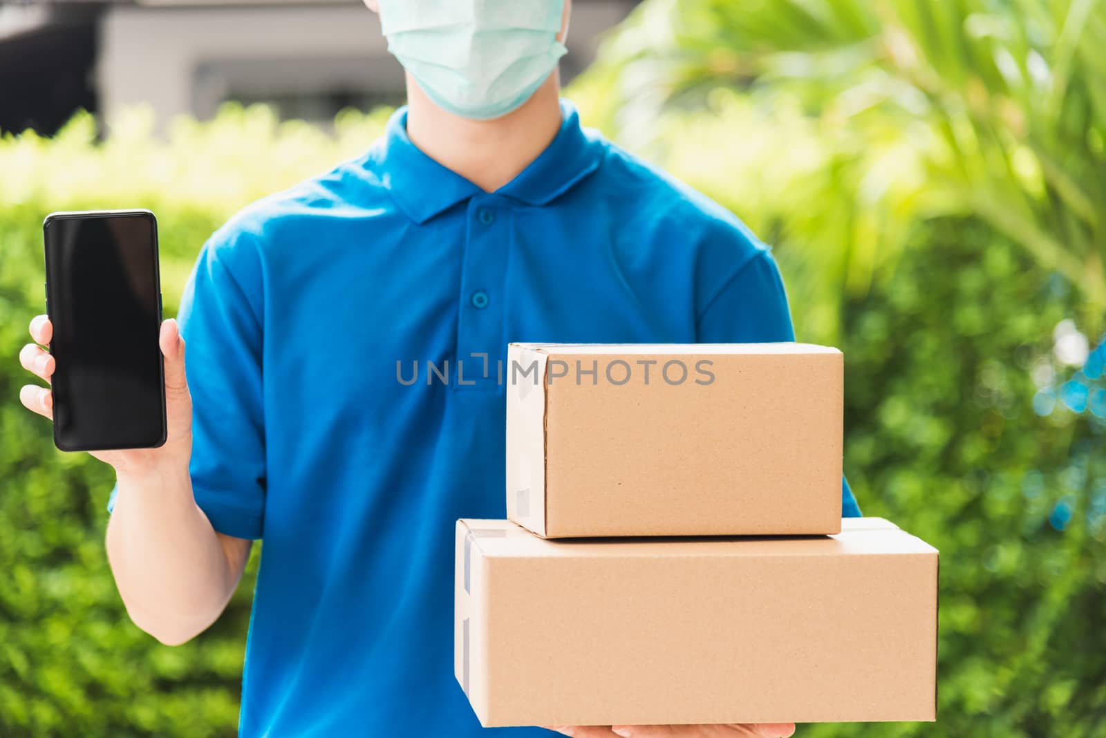 Asian delivery express courier young man giving boxes to customer he wears protective face mask at front home and show mobile phone blank screen, under curfew quarantine pandemic coronavirus COVID-19