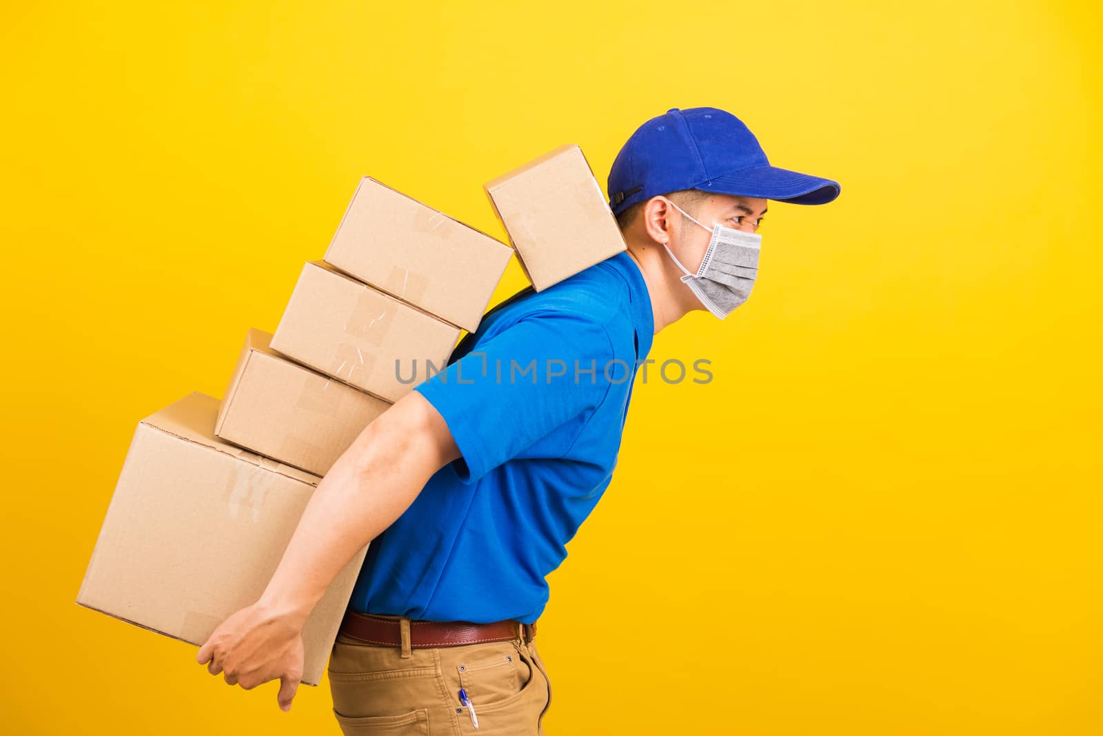 Asian young delivery worker man in uniform wearing face mask protective he has many job lifting stack heavy a lot of boxes on back, under coronavirus COVID-19, studio shot isolated yellow background