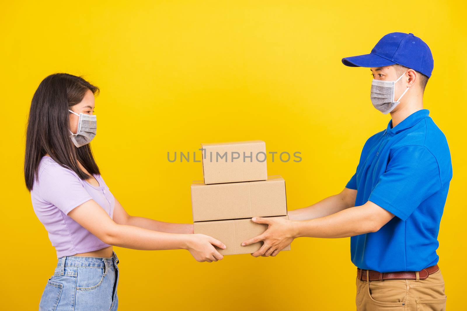 Asian beautiful woman and handsome man wearing protection face mask against coronavirus, accepting delivery boxes, studio shot isolated on yellow background, COVID-19 or corona virus concept